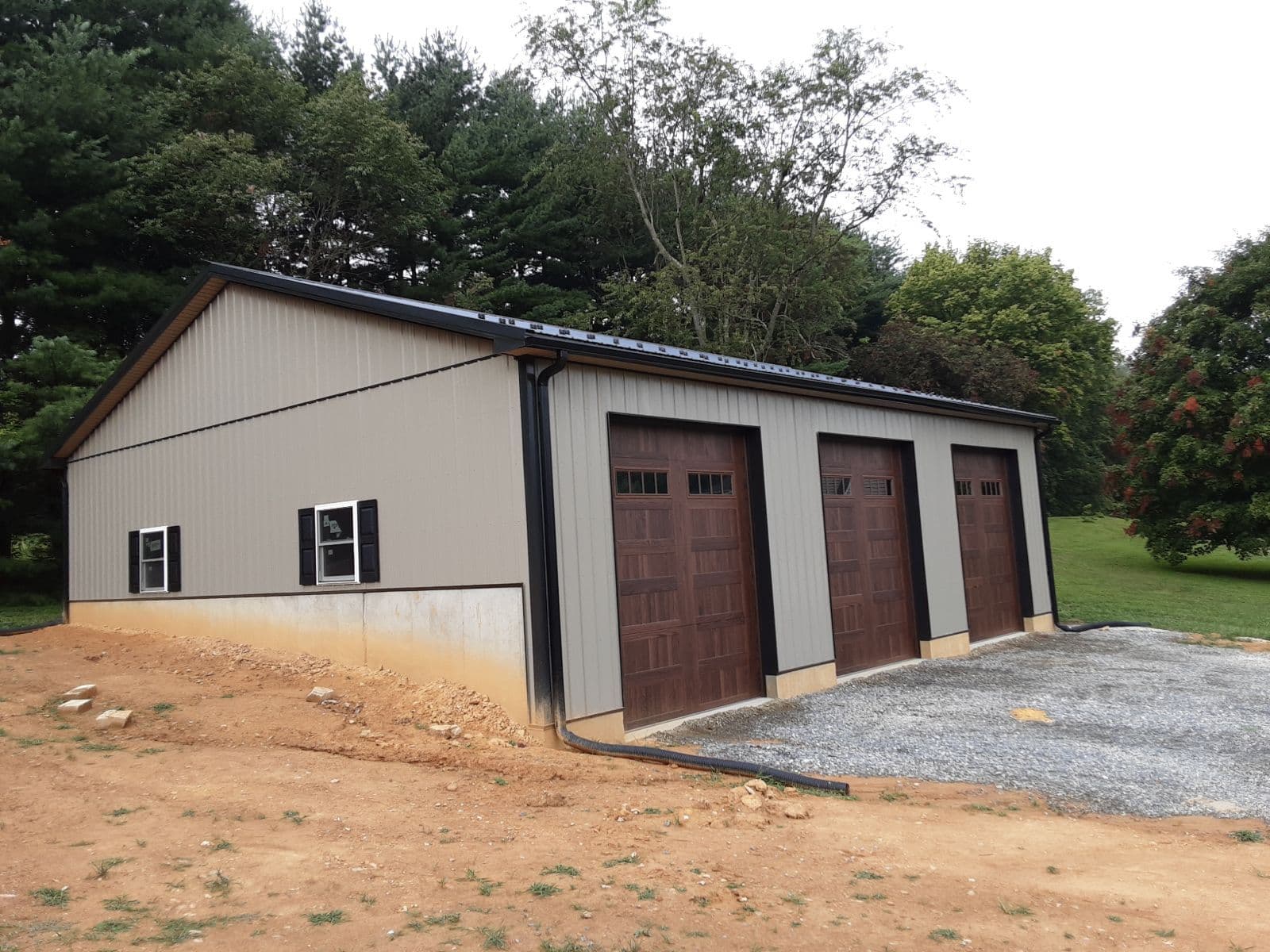 A light gray pole building with black metal roof and trim and three large dark brown garage doors.