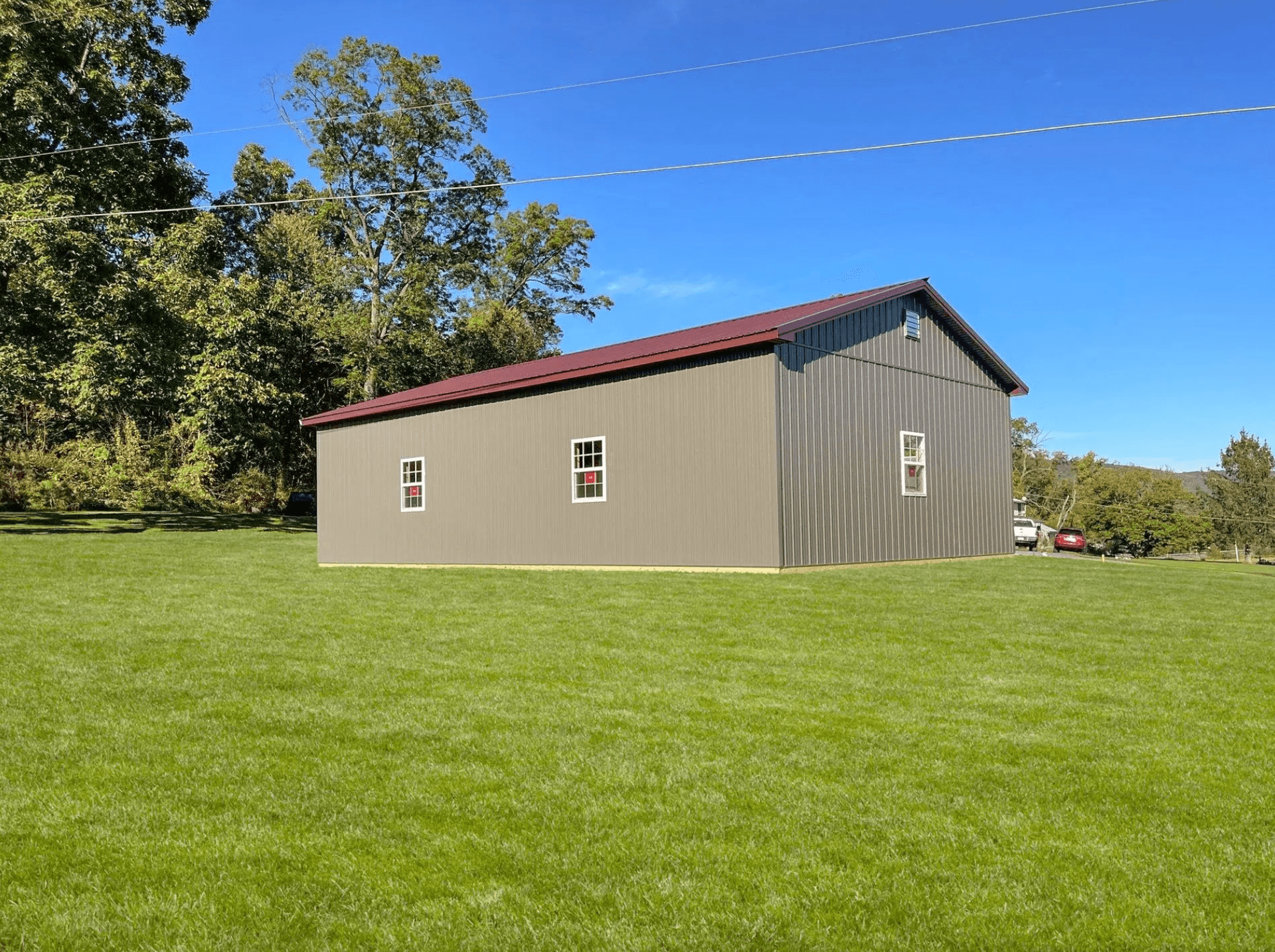 Large green post-frame building with dark metal siding, red metal roof, and white-trimmed windows on a grassy property.