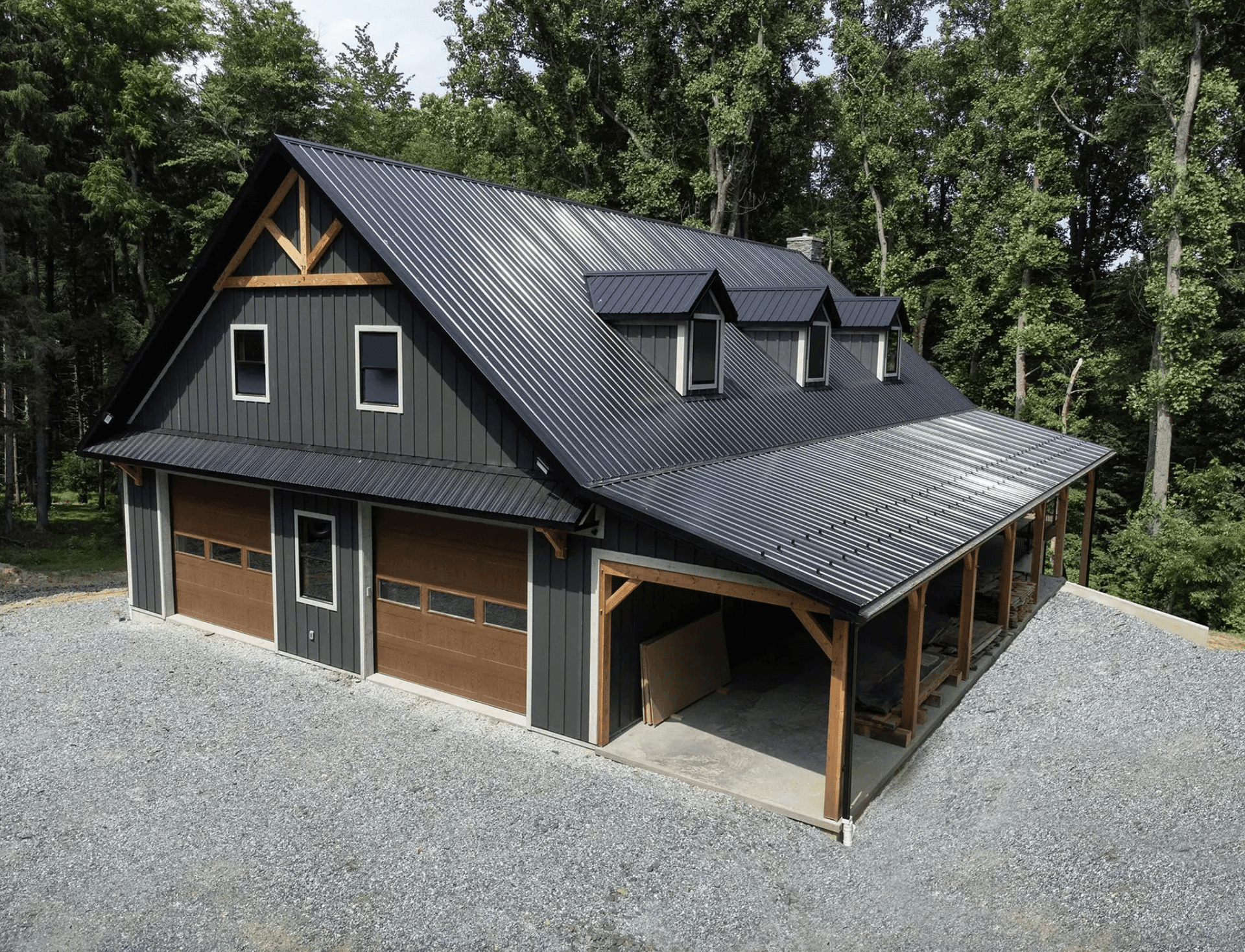 Dark gray post-frame barn with black metal roofing, wooden garage doors, dormer windows, and a covered side porch surrounded by trees
