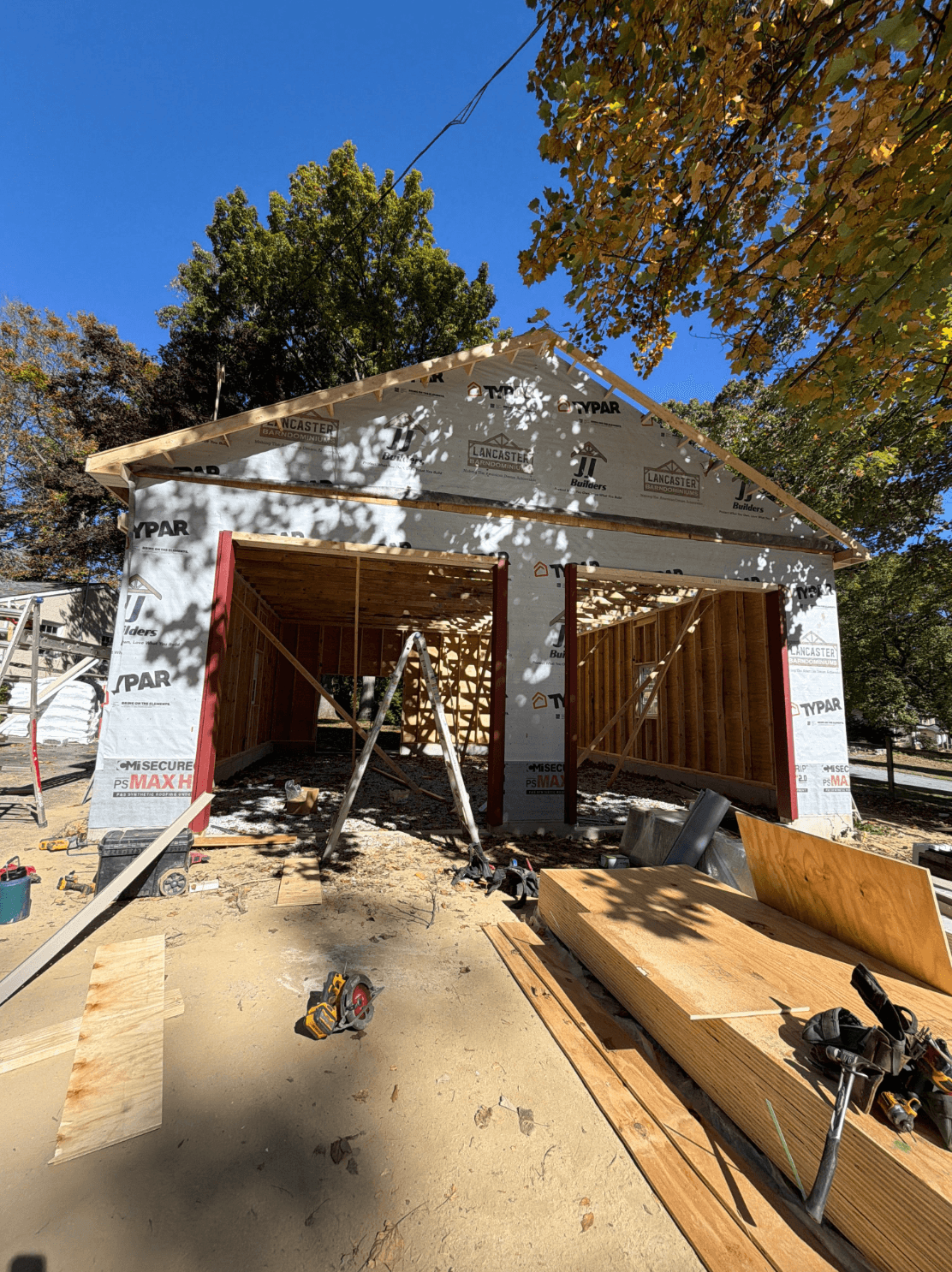 Pole barn under construction with exposed wooden framing, Typar house wrap, open garage bays, and construction materials and tools on site beneath autumn trees.