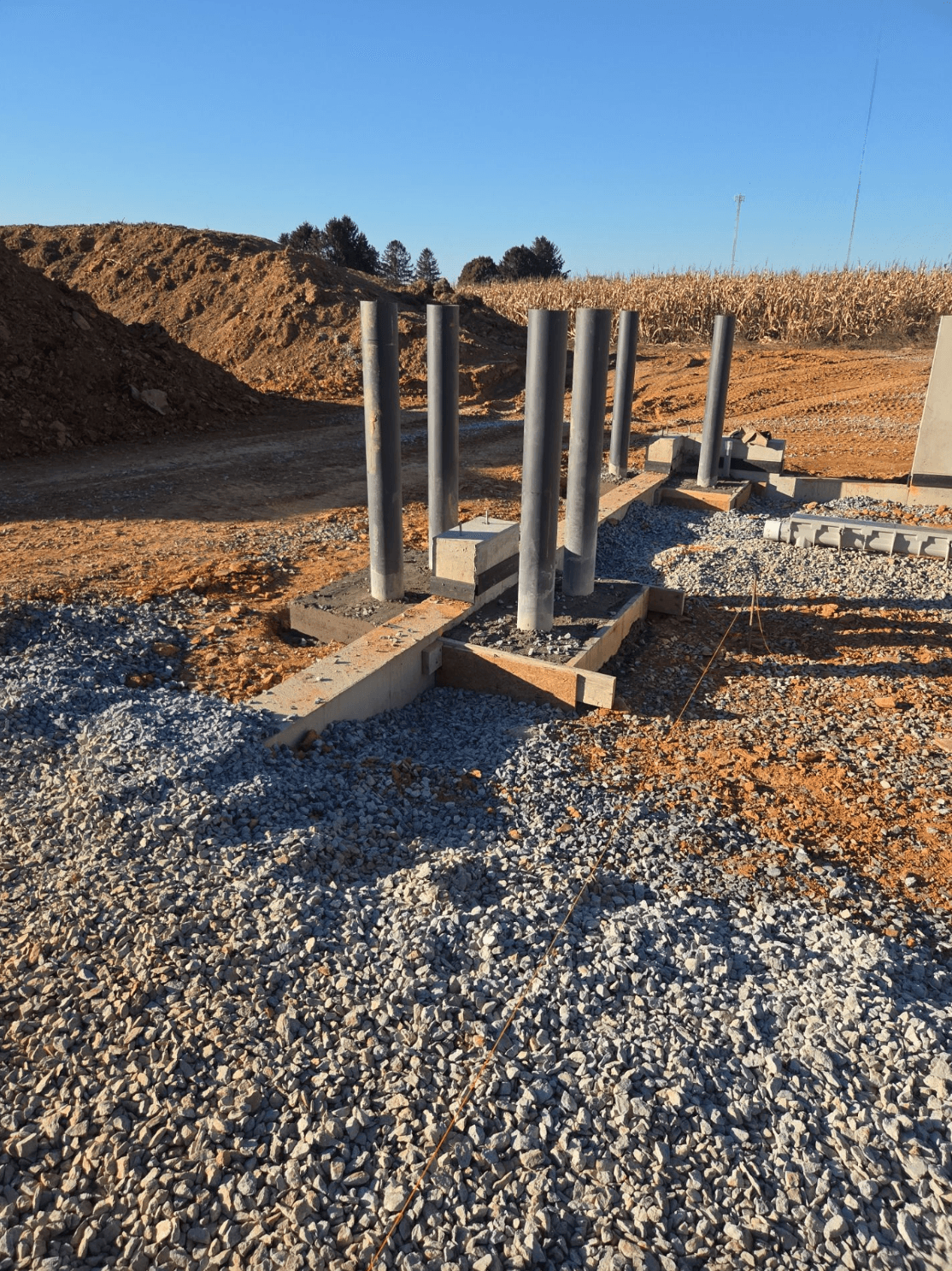 Pole barn construction site showing concrete foundation footings, vertical gray support posts, gravel base, and graded earth beside a harvested cornfield