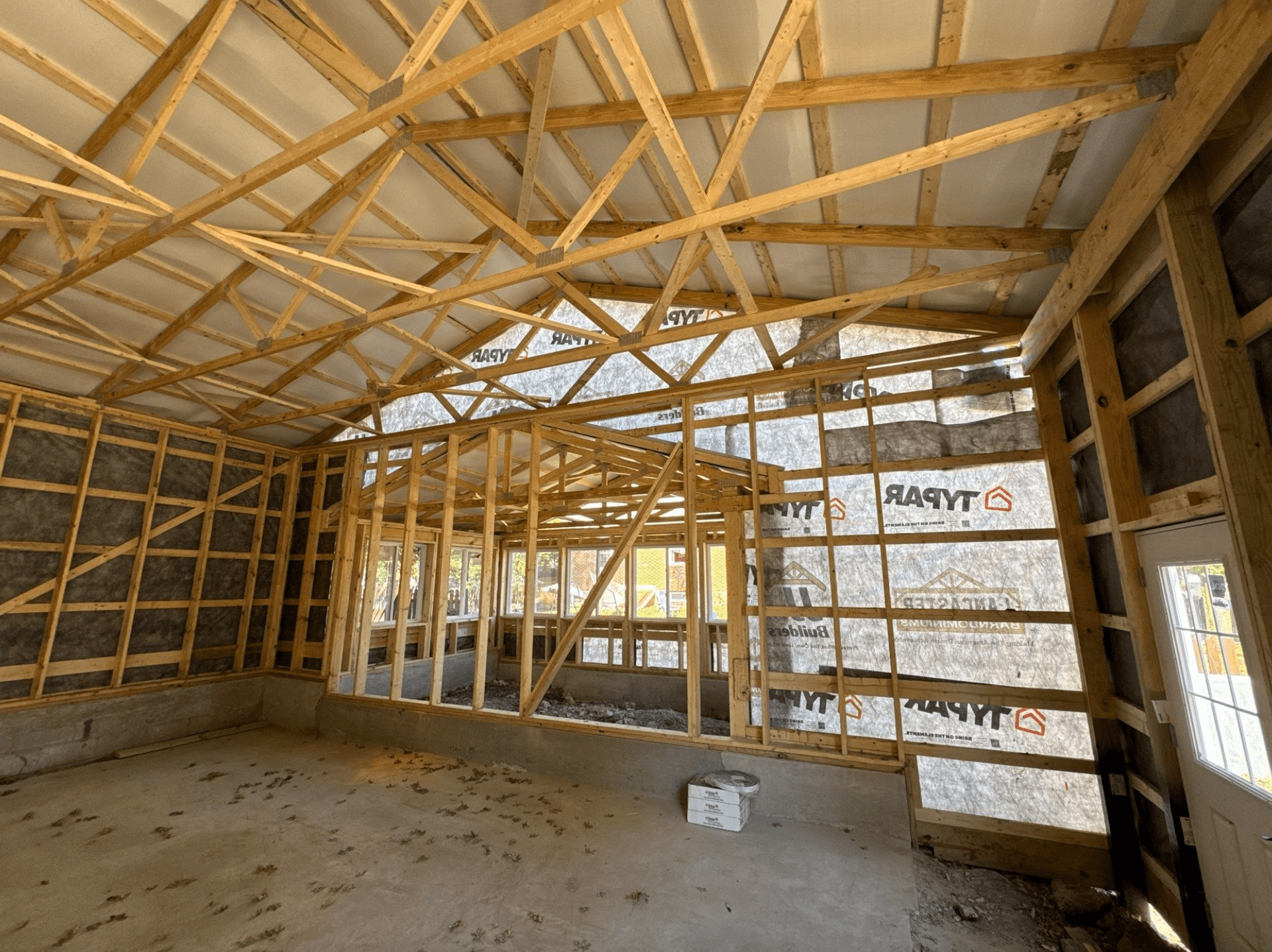 Interior of a post-frame building under construction, featuring exposed wooden roof trusses, wall framing, house wrap, insulation, and a concrete floor.
