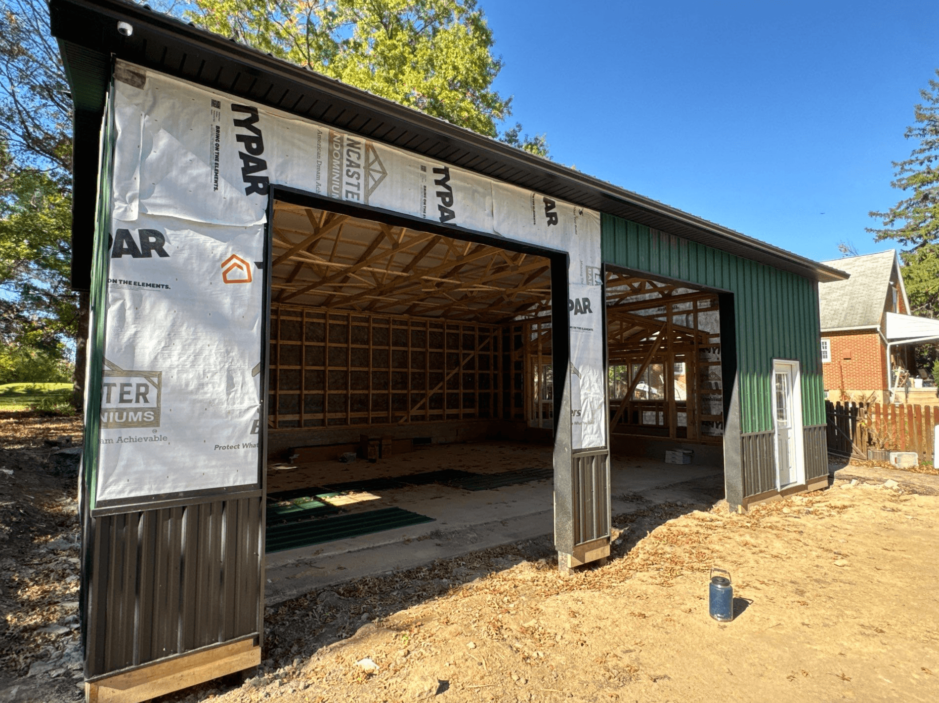 Green post-frame building under construction with large open garage bays, exposed wooden roof trusses, metal siding, and protective house wrap around the front openings.