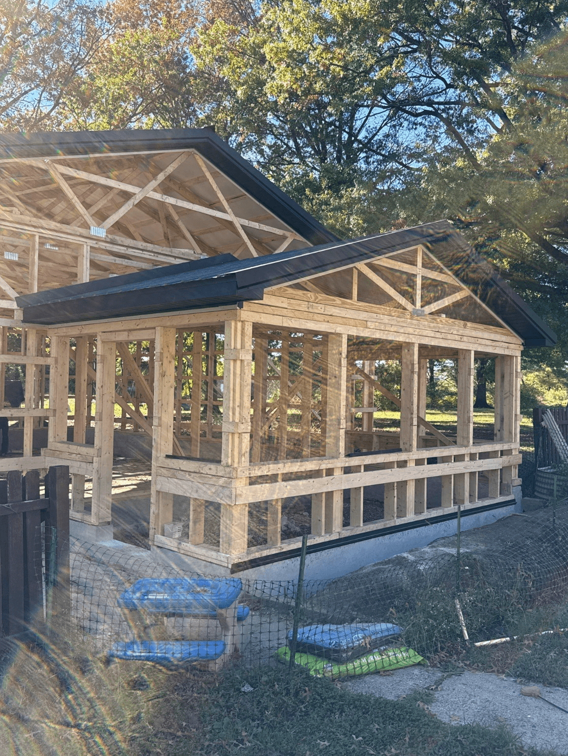 Custom post-frame building under construction with exposed wooden framing, open wall sections, black metal roofing, and a concrete foundation surrounded by trees