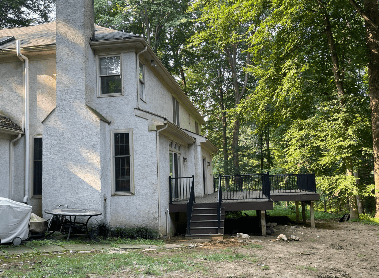 White stucco home with a raised wooden deck and stairs surrounded by mature trees.