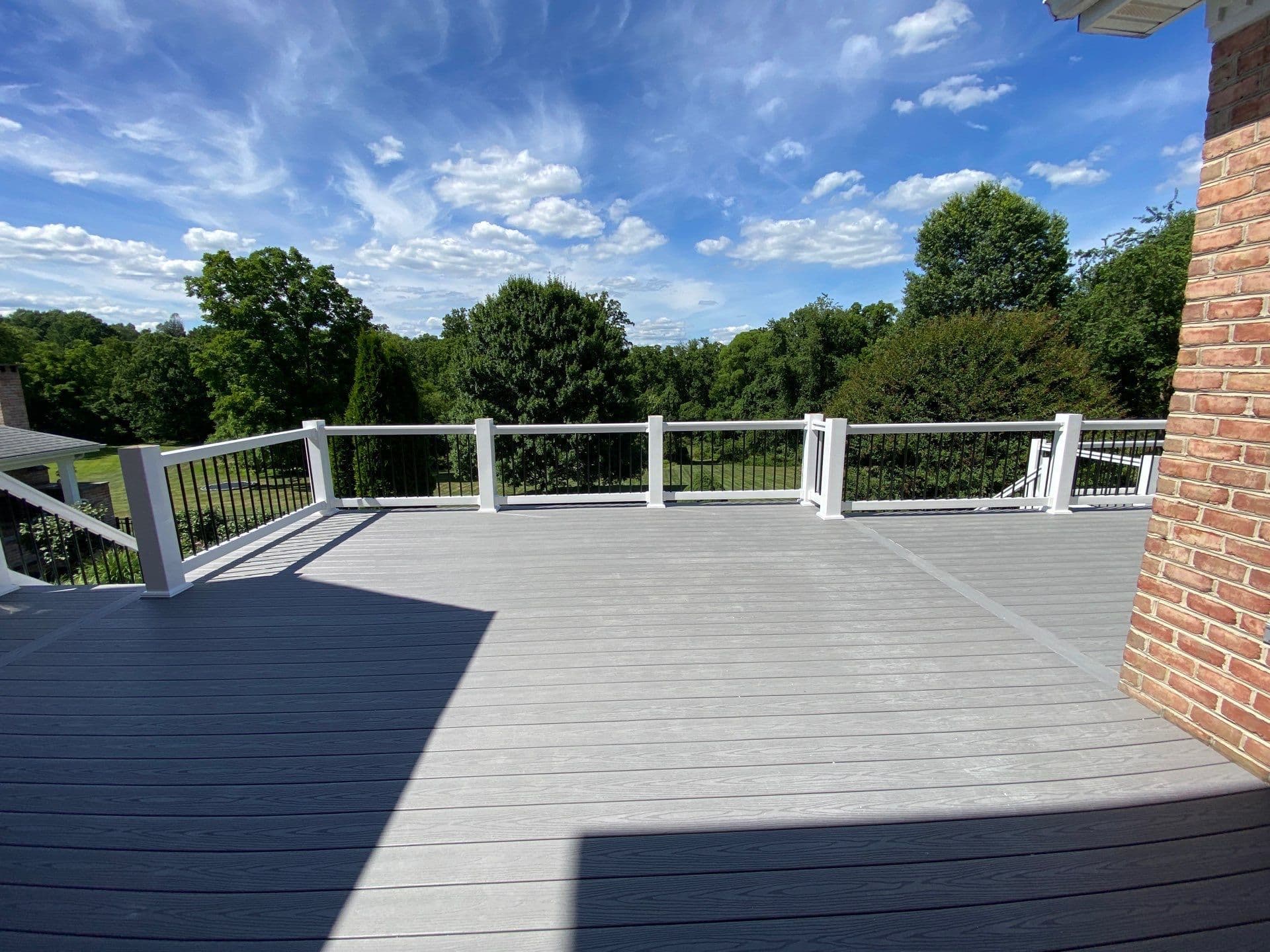 Crafted by custom builders, this spacious outdoor deck boasts gray flooring and a white railing, offering a breathtaking view of lush green trees under a partly cloudy sky.