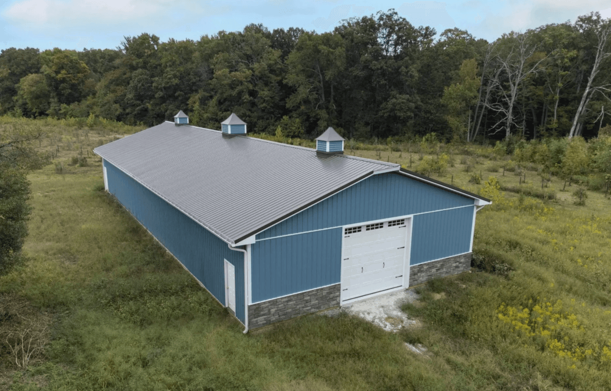 Blue metal building with a large garage door surrounded by grassy fields and trees.
