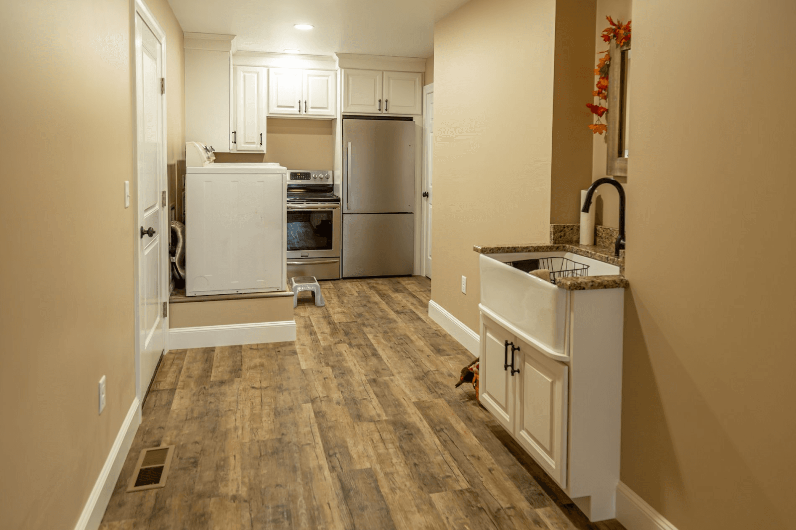 Narrow laundry room with wood flooring, white cabinets, a farmhouse sink, and appliances including a washing machine and fridge. Beige walls are adorned with a floral decoration.