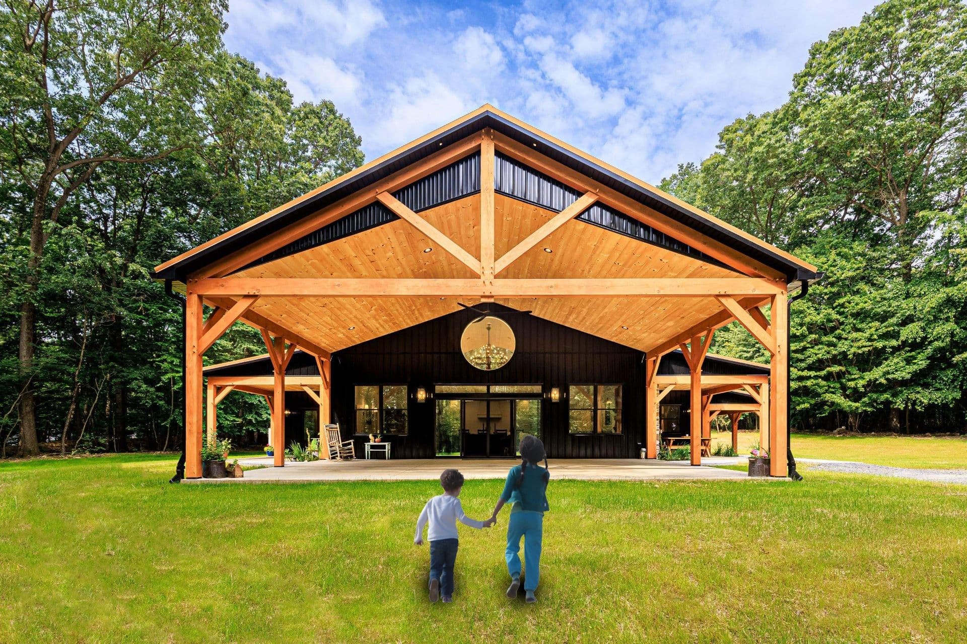 Two children hold hands in front of a new black and brown barndominium with a large covered area.