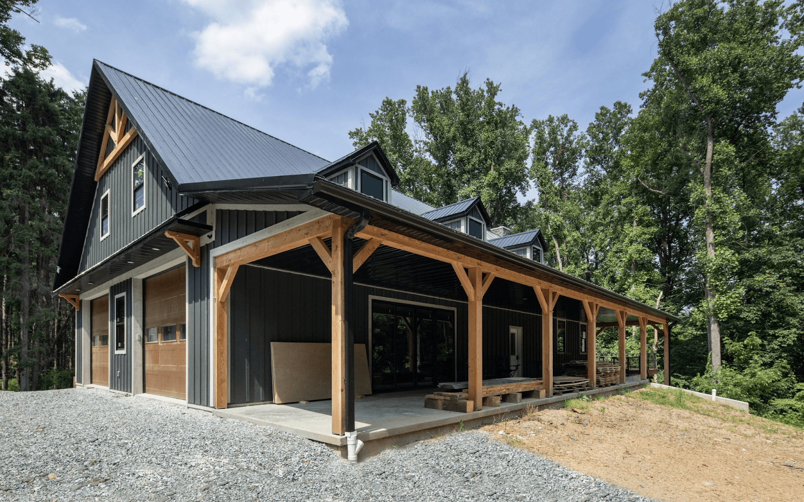 A black post frame building with a black roof, porch, garage bays, and brown accents.