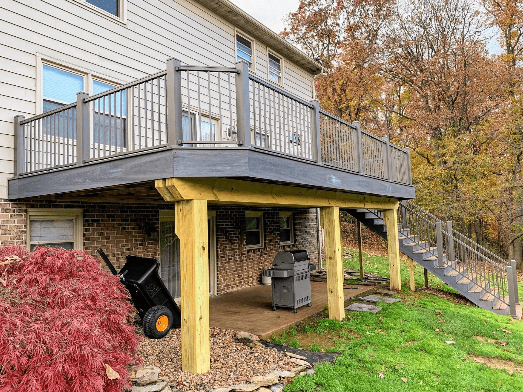 Elevated backyard deck with gray railings, wood posts, stairs, and patio space beneath a two-story home.