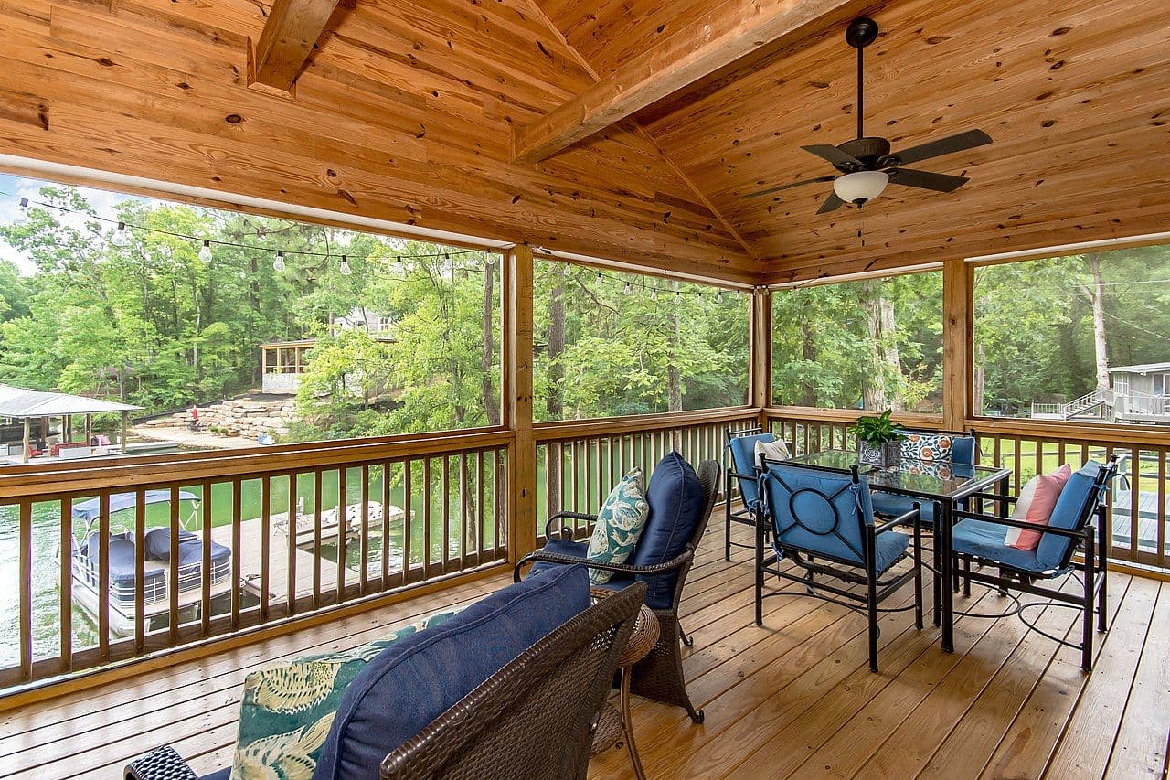 A covered deck with patio furniture overlooks a lake.