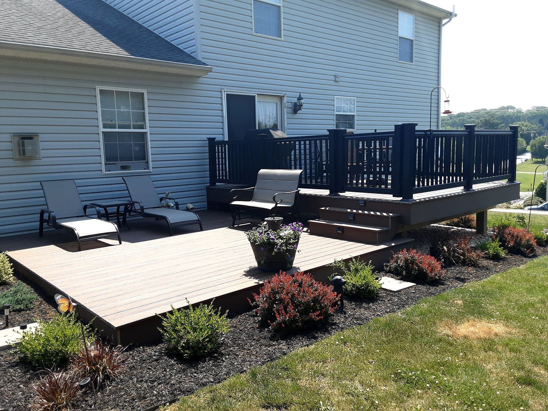 A backyard oasis featuring a deck with lounge chairs and potted plants, skillfully crafted by custom builders. Adjacent to this is a raised wooden deck with railings, perfectly complementing the two-story house with siding.