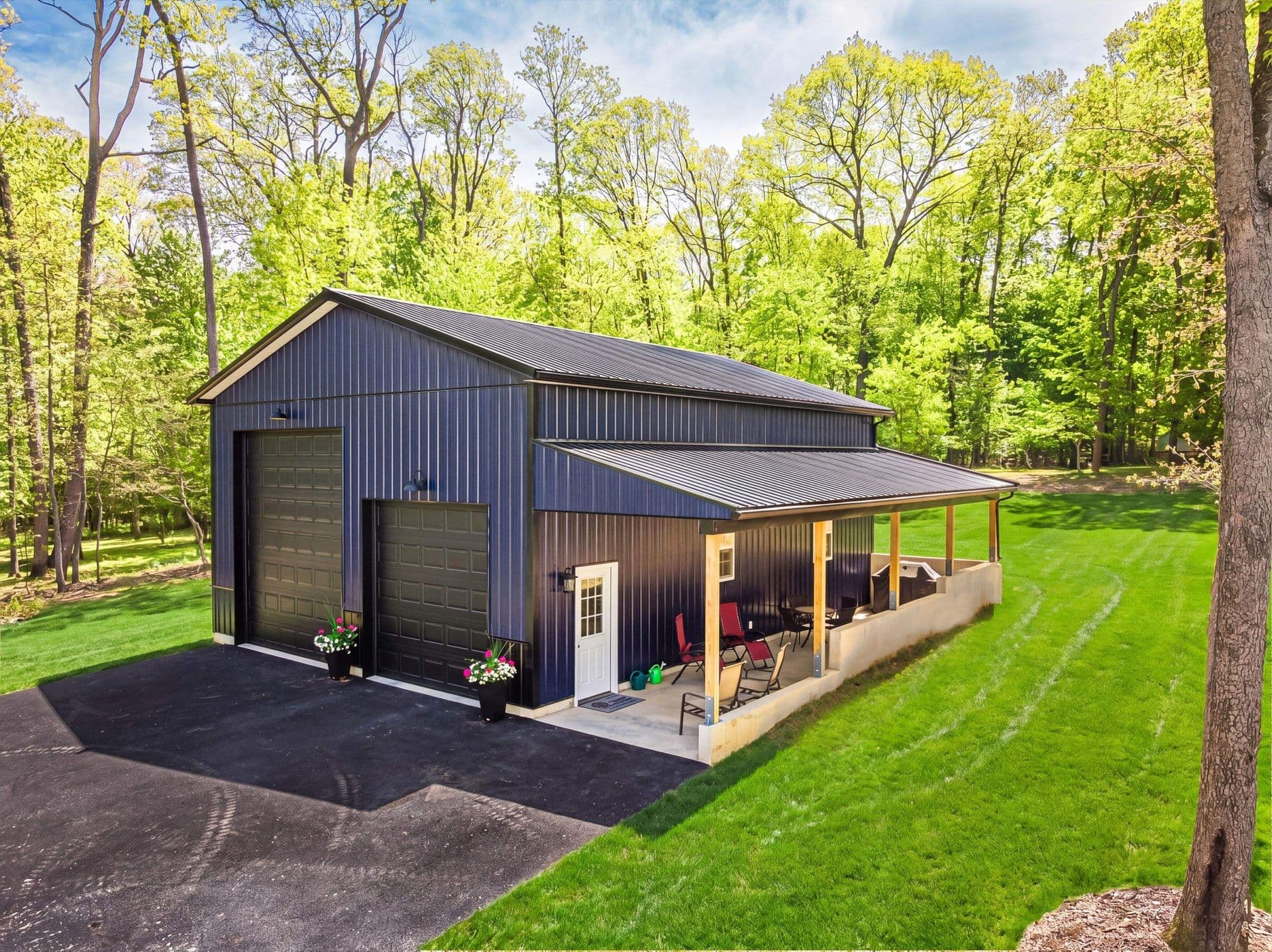 Dark navy metal pole barn with covered porch, double garage doors, and patio seating surrounded by green trees