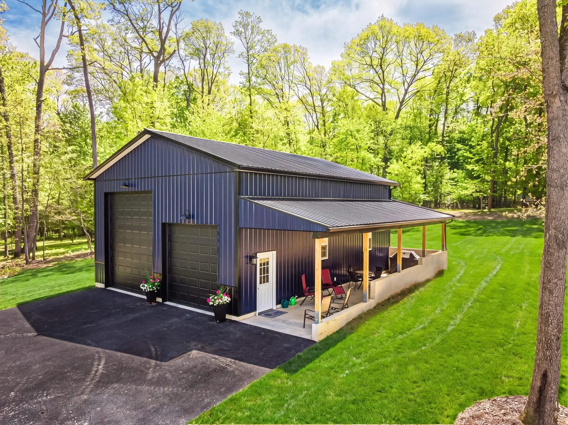 Dark navy metal pole barn with covered porch, double garage doors, and patio seating surrounded by green trees