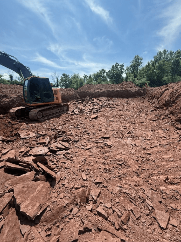 Excavator working on a rocky construction site with exposed soil and rock under a clear blue sky.