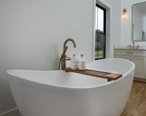 A modern bathroom with a white freestanding tub, bronze faucet, and wooden tray holding two bottles. A large mirror and sink are visible in the background. Light wood flooring.