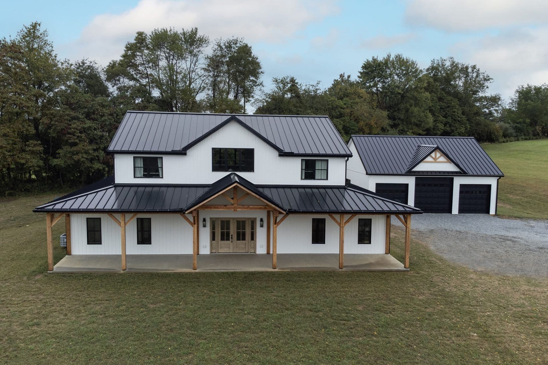 Two-story white house with black metal roof, front porch with wooden beams, and detached two-car garage on a grassy property with trees in the background.