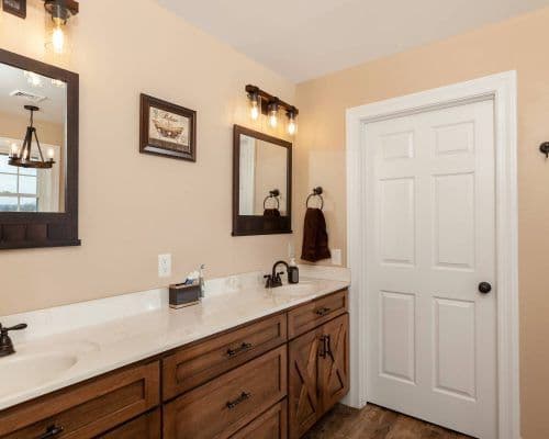 Bathroom interior with dual sinks, wooden cabinetry, two mirrors, and light fixtures. Beige walls and a white door are visible. A small framed picture and towel hang on the wall.