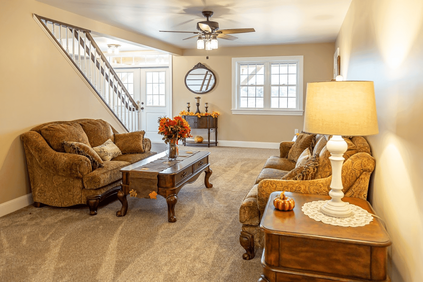 A cozy living room with two brown couches, a wooden coffee table with autumn decorations, a lamp, and a staircase in the background. A mirror and window are on the far wall.