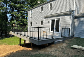 A gray raised deck with black metal railings, crafted by custom builders, is attached to a two-story house. A sliding glass door opens to the deck, while trees and grass provide a serene backdrop.