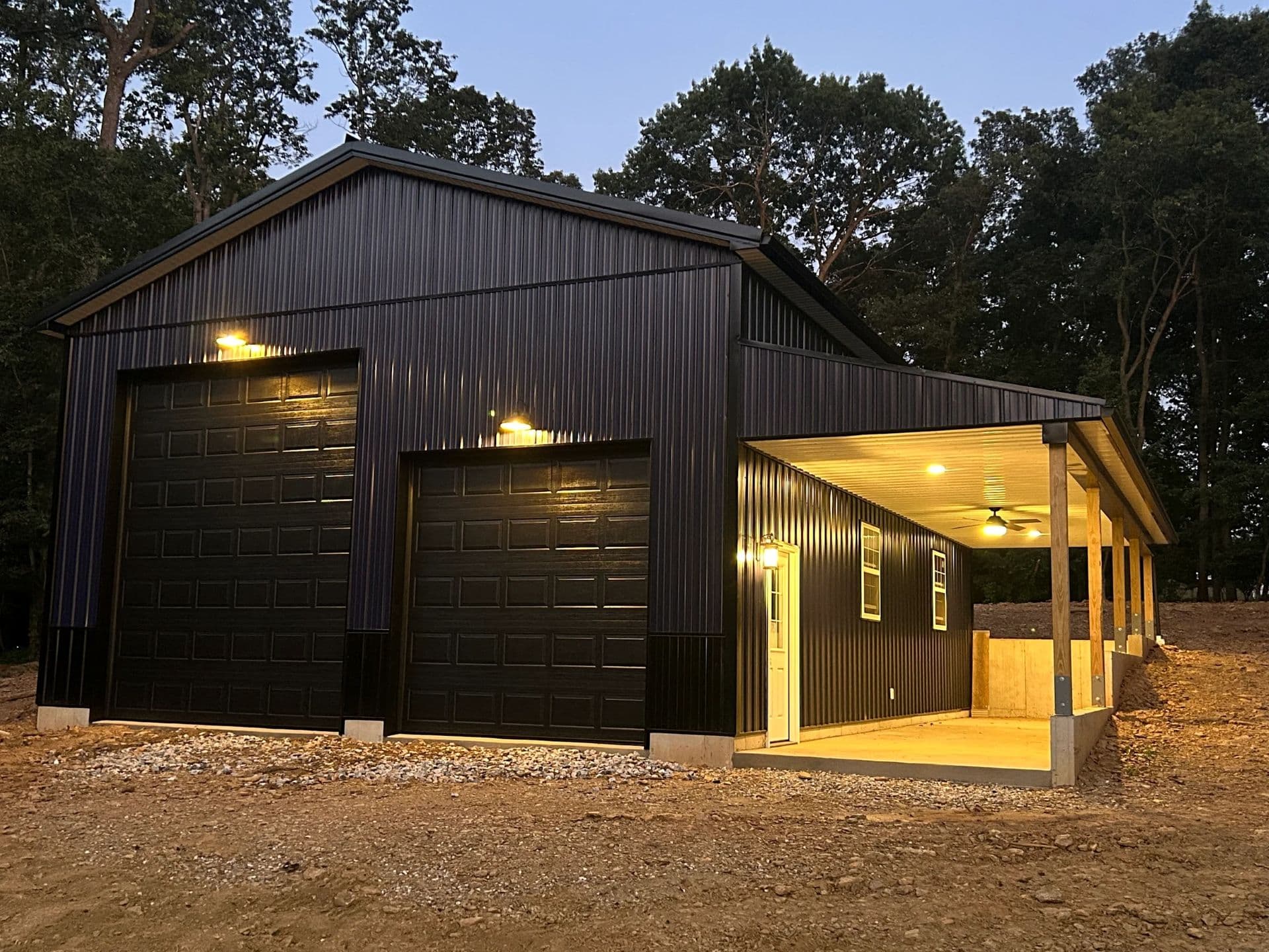 A newly-built black post frame garage with black garage doors lit up at dusk.