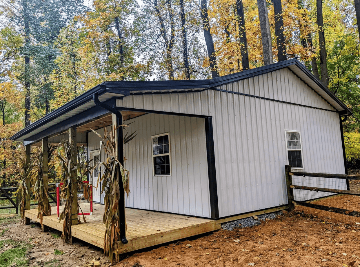 White metal-sided barn with a covered wooden porch surrounded by trees and autumn foliage.
