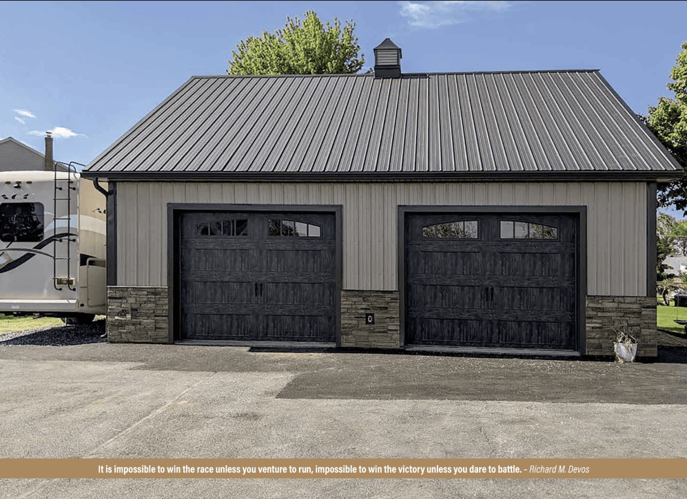 A tan post frame building with two garage bays with black doors and faux stone wainscoting.